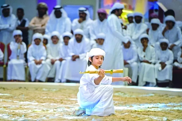 Emirati child performing Al-Ayyala (Yowlah) with a bamboo stick in traditional white dress, with an audience of Emirati men and children watching (2014) — representing the intergenerational transmission of the traditional performing art inscribed on UNESCO's Representative List as Intangible Cultural Heritage File 01012 by the UAE and Oman