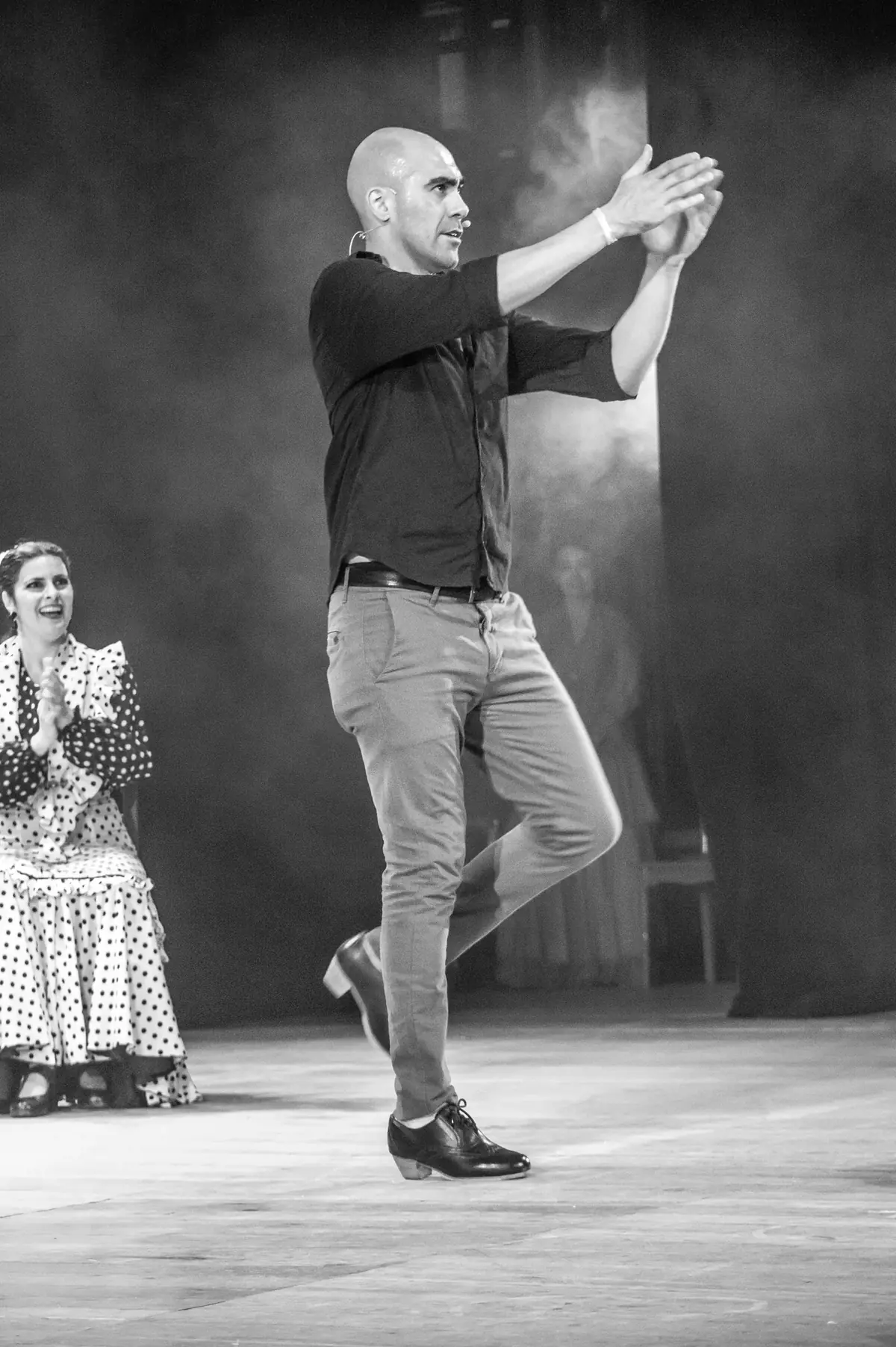 Male flamenco bailaor performing zapateado (footwork) mid-step on stage with female dancer doing palmas (hand-clapping) in background, Spain — demonstrating the baile (dance) element of flamenco inscribed on UNESCO's Representative List of the Intangible Cultural Heritage of Humanity as File 00363