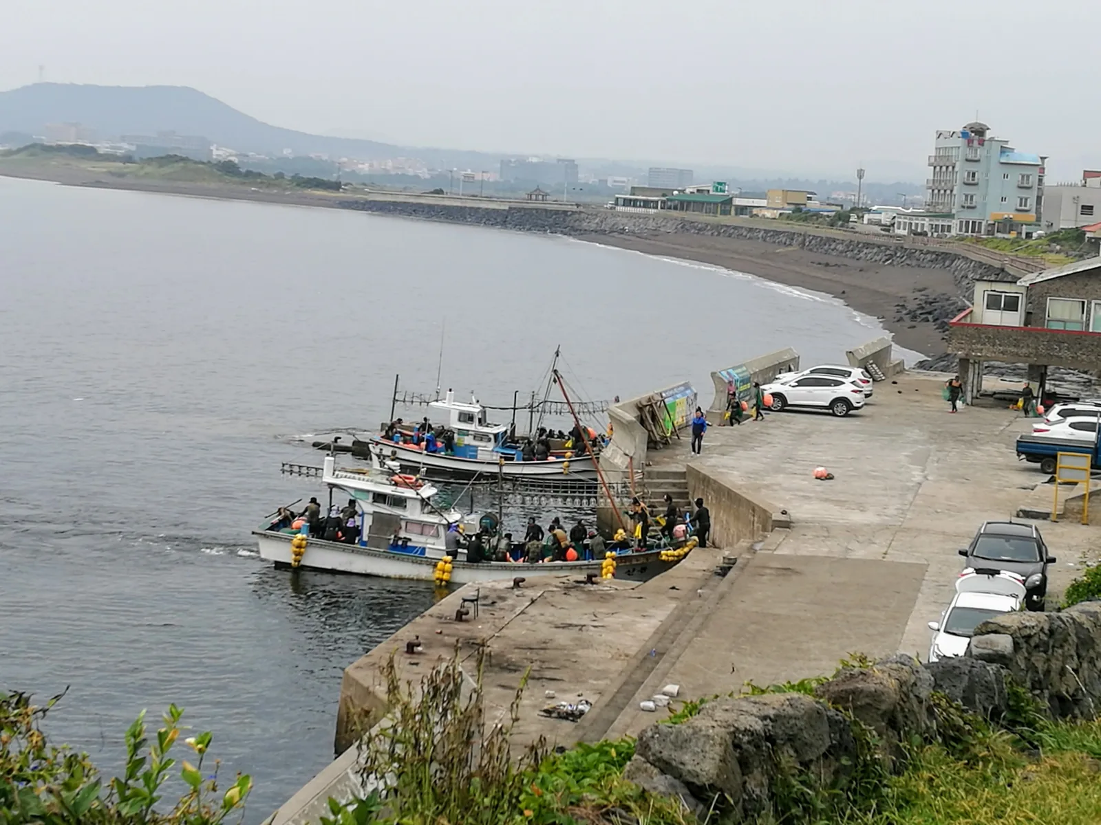 Haenyeo women divers in wetsuits boarding fishing boats at Jeju Island harbor with Seongsan volcanic peak visible — Culture of Jeju Haenyeo (File 01068) was inscribed on UNESCO's Representative List in 2016, recognizing the tradition of Jeju's female free-divers who harvest seafood without breathing apparatus