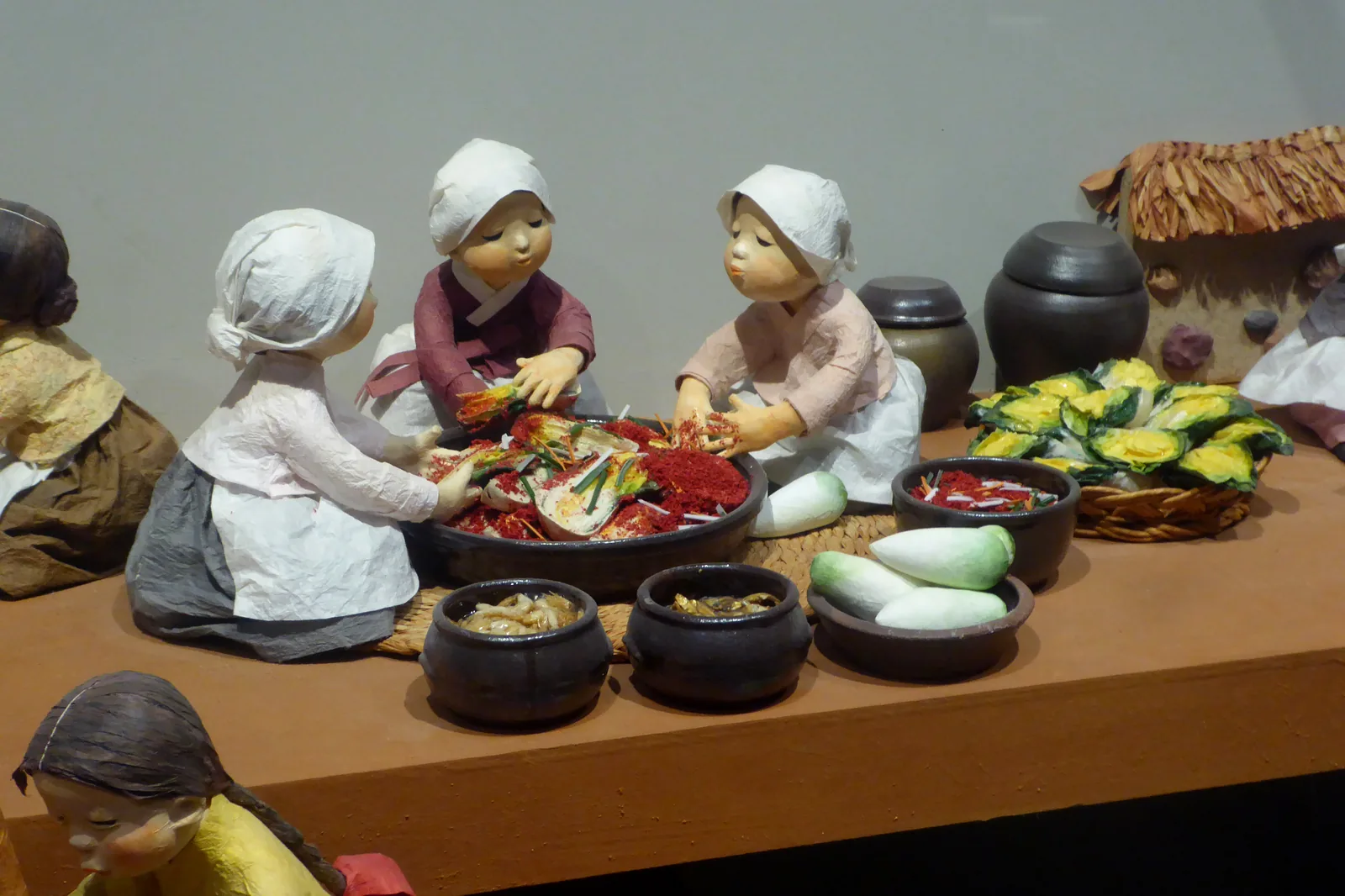 A museum diorama at the National Folk Museum of Korea in Seoul depicting women in traditional hanbok applying chili seasoning to cabbage leaves in a large bowl — with onggi earthenware jars and vegetables visible — illustrating the traditional kimjang process (UNESCO ICH File 00881) as practiced in Joseon-era Korean households