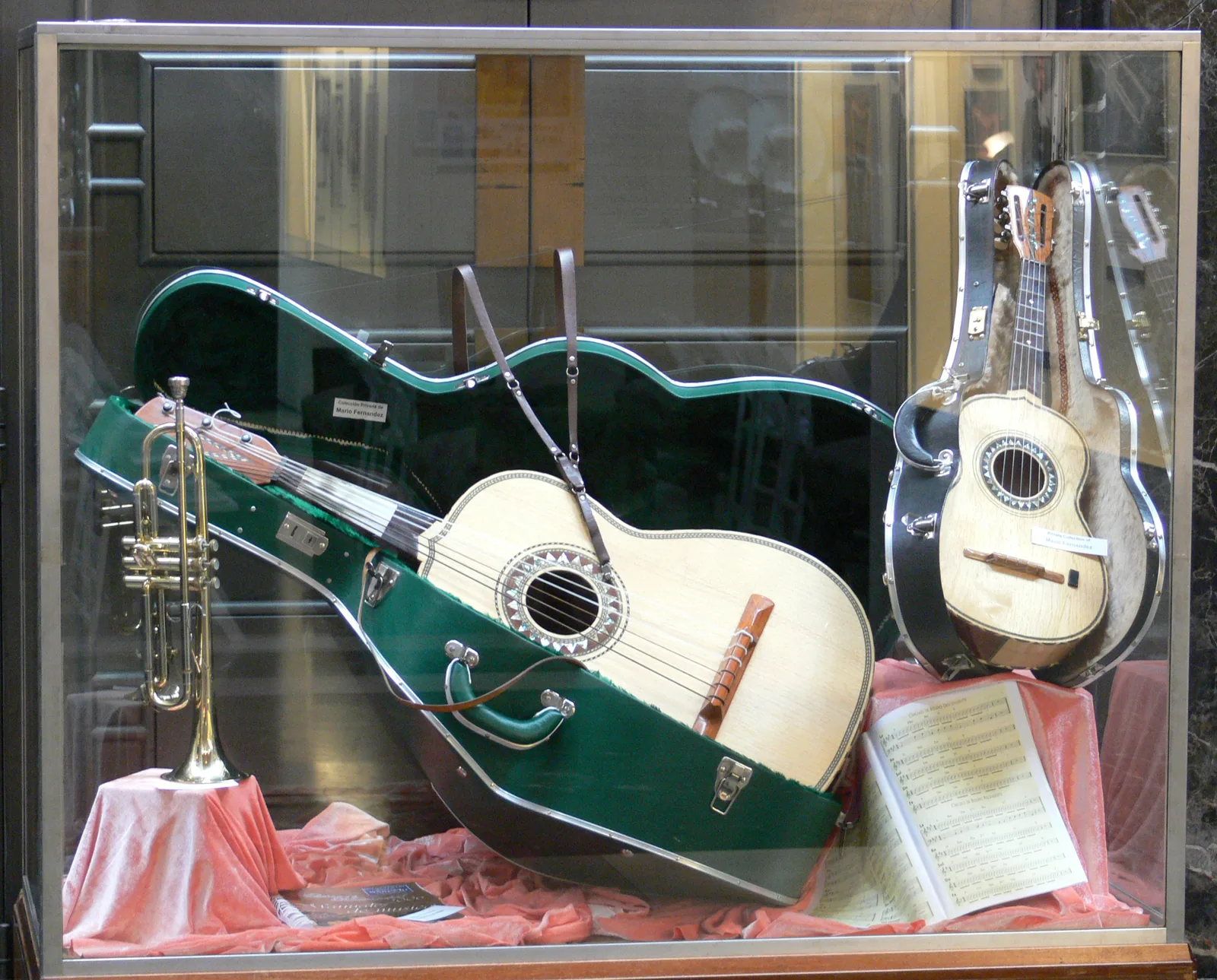 Mariachi instruments in a museum display case showing the guitarrón (large fretless acoustic bass in green case), vihuela (five-string rhythm guitar with convex back, right), and trumpet — the guitarrón and vihuela are the sonic fingerprints that distinguish mariachi from other regional Mexican styles recognized in UNESCO's 2011 ICH inscription