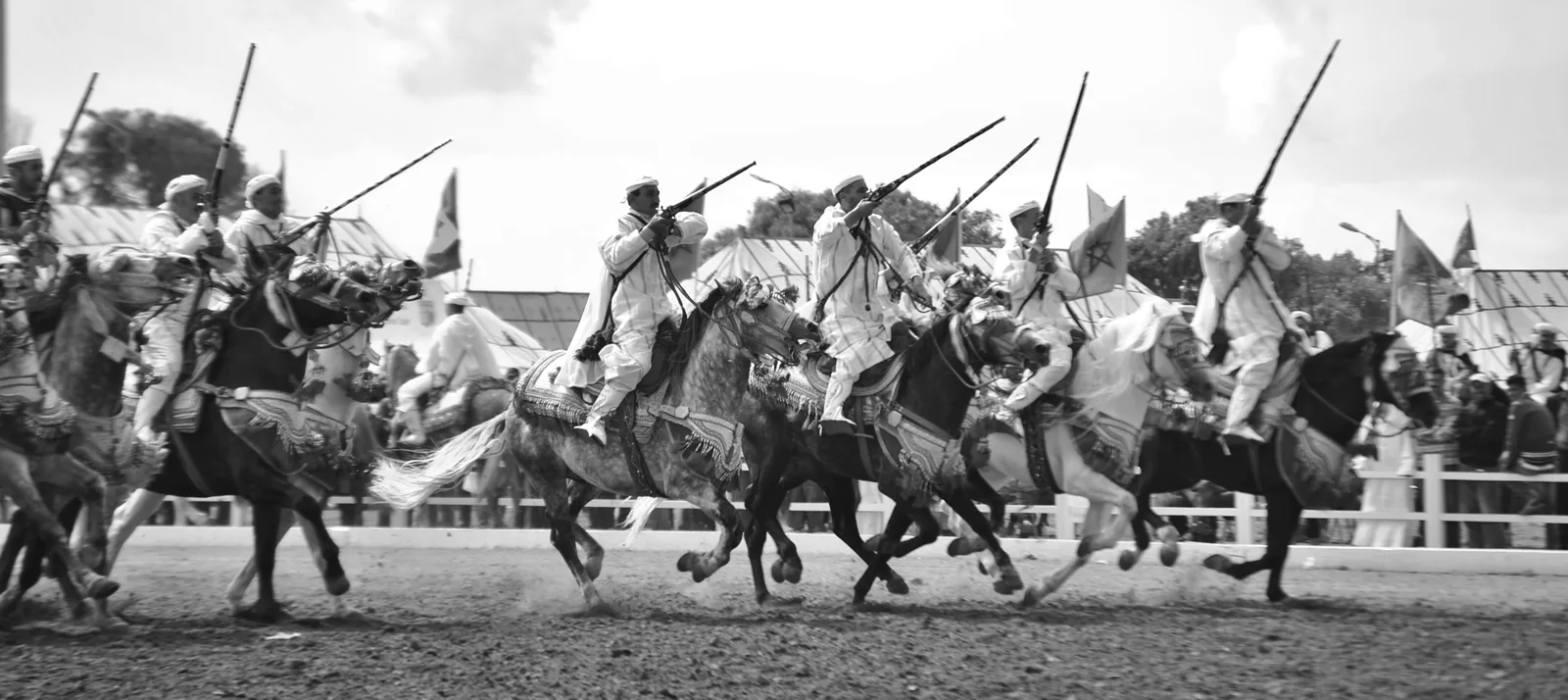 Tbourida riders in white traditional dress charging in precise formation with muskets raised at the Horse Festival in El Jadida, Morocco — the UNESCO-inscribed equestrian performance (File 01483, 2021) in which riders charge simultaneously and fire at the end of the run in a simulation of historical cavalry formation