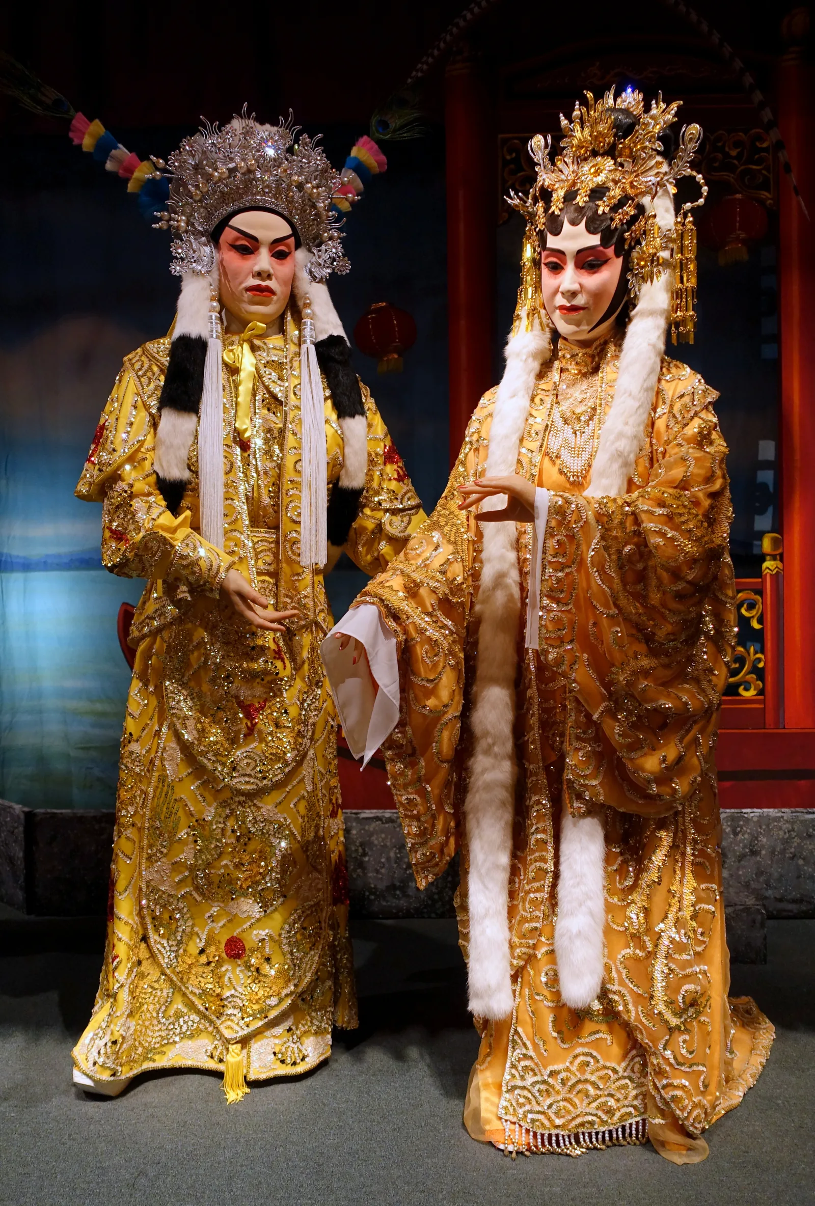 Two traditional Chinese opera figures in gold embroidered imperial court costumes with elaborate headdresses and water sleeves at the Hong Kong Museum of History — the flamboyant embroidered silk costumes and pearl headdresses of Peking opera encode rank, role type, and dynasty in a symbolic theatrical language