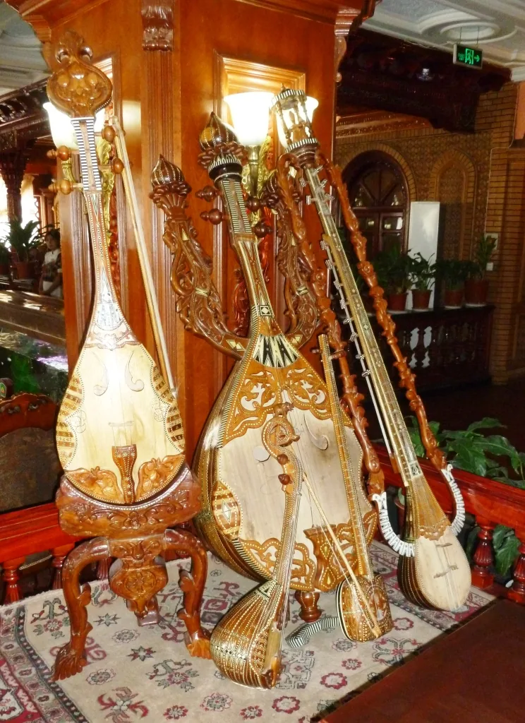 Traditional Uyghur musical instruments displayed at a Kashgar restaurant — multiple ornately decorated rawap and dutar instruments with carved wooden bodies and inlay work, representing the principal plucked string instruments of the Uyghur Muqam of Xinjiang (UNESCO ICH File 00109)