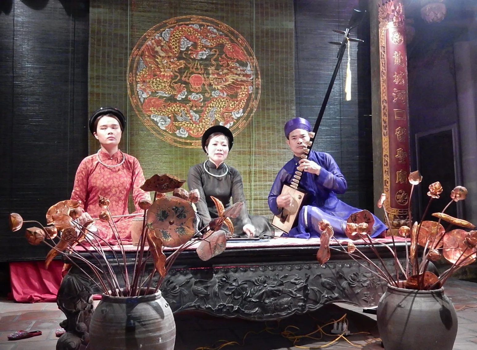 Vietnamese Ca Trù chamber music performance in Hanoi (2015): female ca nương singer in black áo dài, female attendant in pink áo dài, and male musician playing đàn nguyệt lute — Ca Trù singing (File 00309) is on UNESCO's List of Intangible Cultural Heritage in Need of Urgent Safeguarding