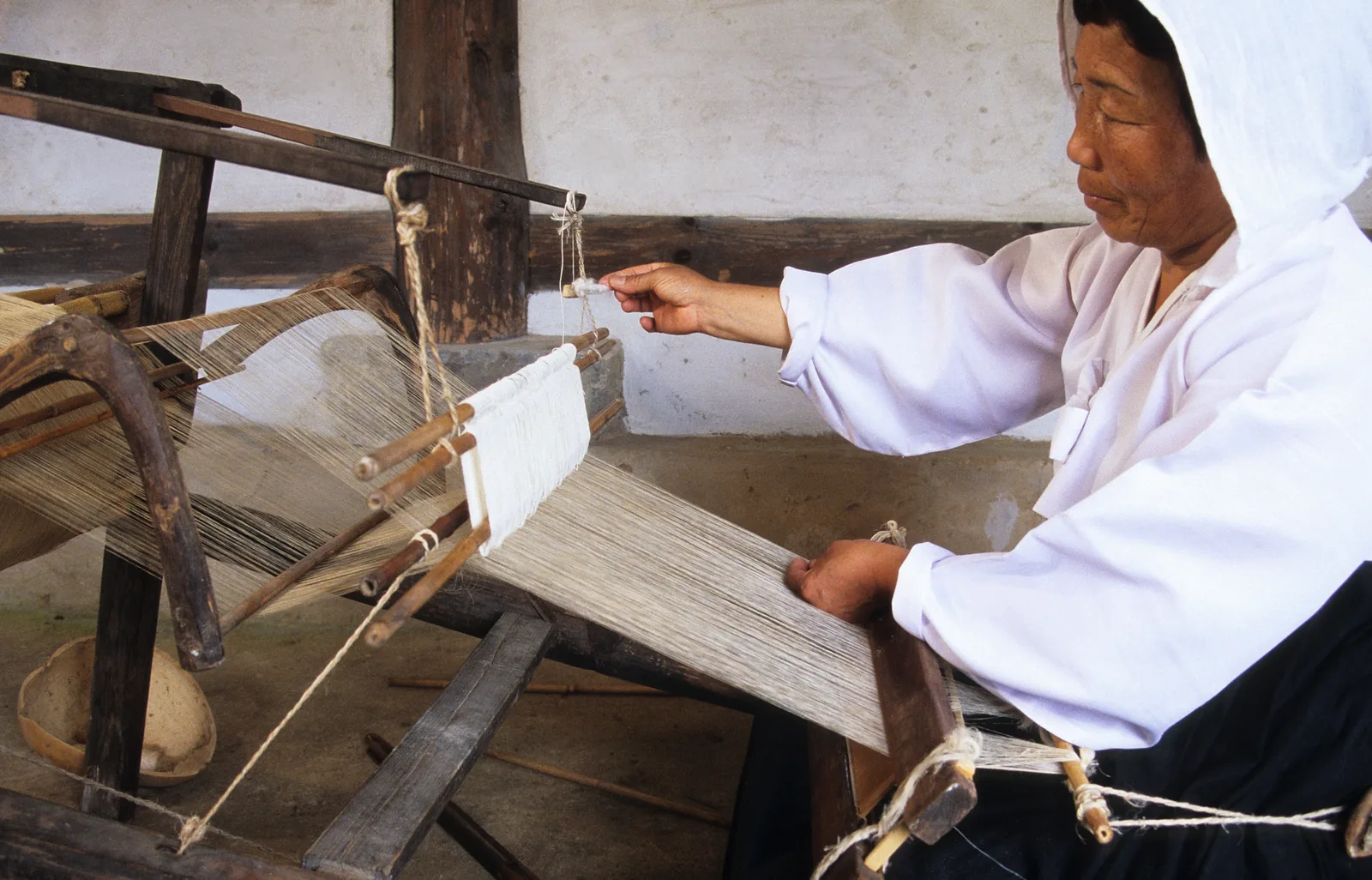 Korean woman at a traditional Hansan mosi loom performing the thread-joining process (실올 잇기) in Seocheon, South Chungcheong Province, 2003 — documented by the National Intangible Cultural Heritage Center showing the ultra-fine white ramie threads and traditional Korean standing loom of the craft inscribed as UNESCO ICH File 00453